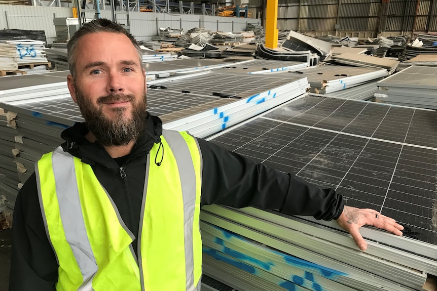 A man in a high-viz vest rests his arm on a stack of old solar panels inside a warehouse.