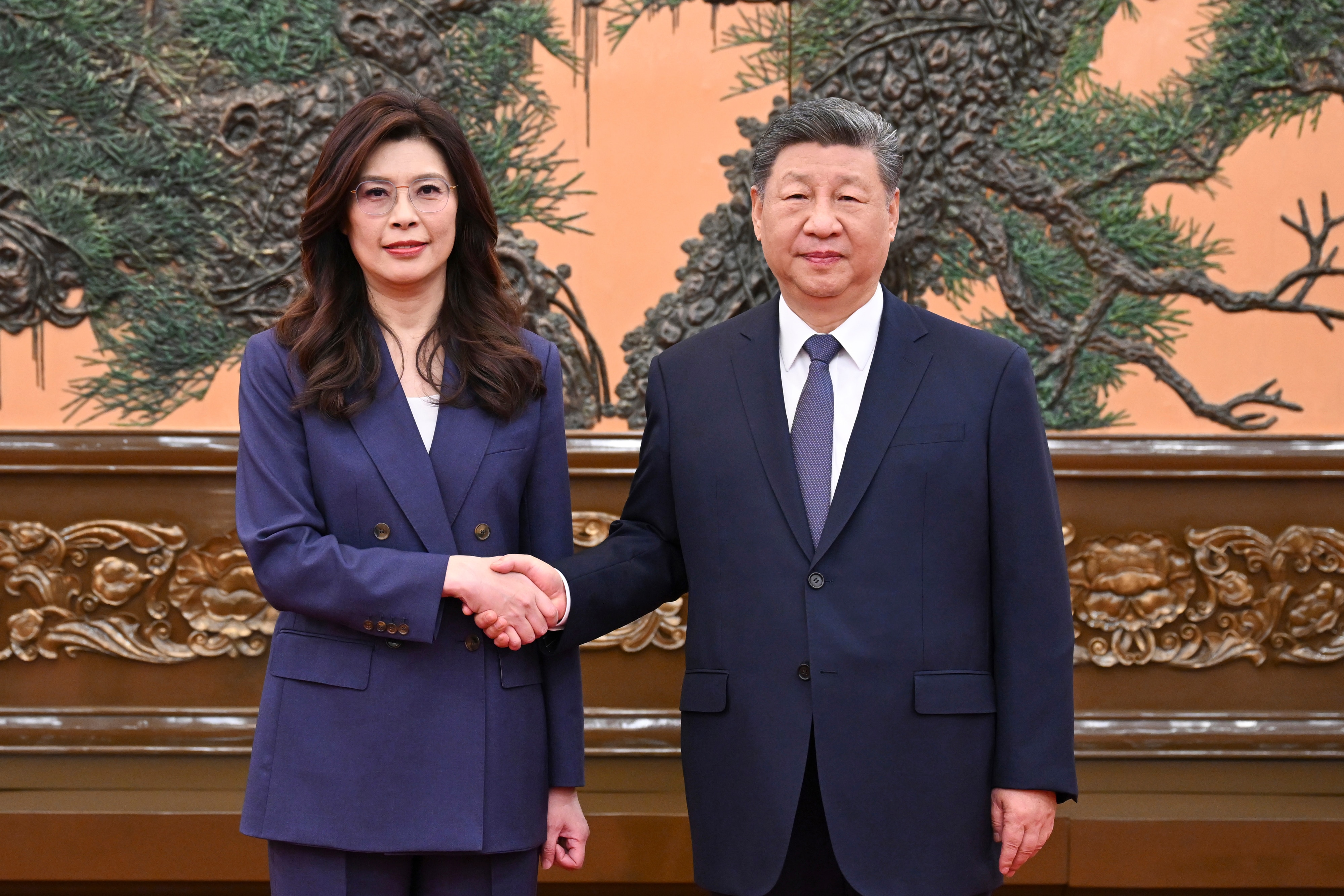 A man and woman, both dressed in suits, shake hands formally as they face the camera.