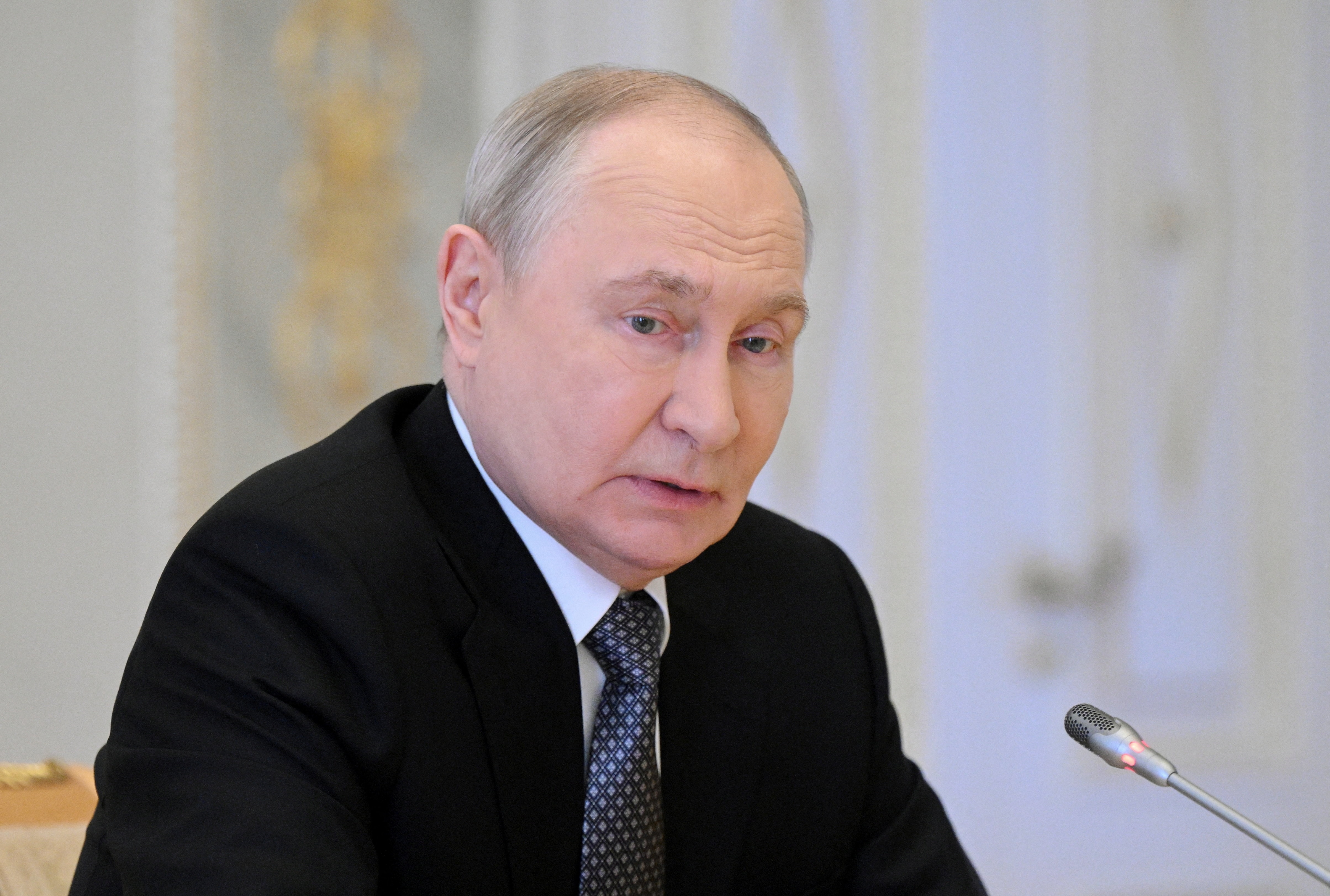 Vladimir Putin sitting at a conference table speaking into a microphone.