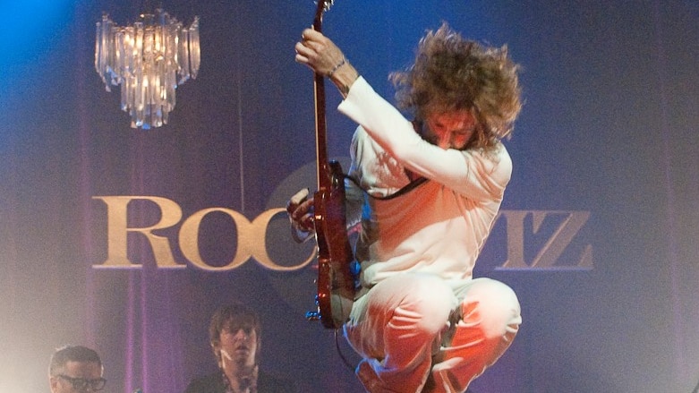 Tim Rogers on stage playing guitar as he leaps into the air
