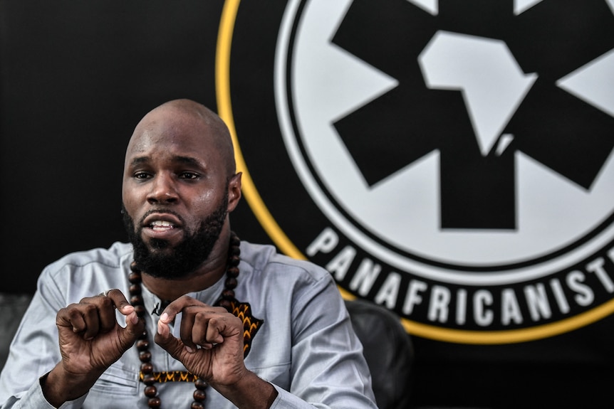 Kemi Seba sits at a press conference with a Pan Africanism banner behind him. 