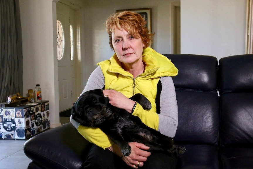 Serious middle-aged woman, short red hair, wears yellow puffer, grey top, black dog in lap, black sofa, front door behind.