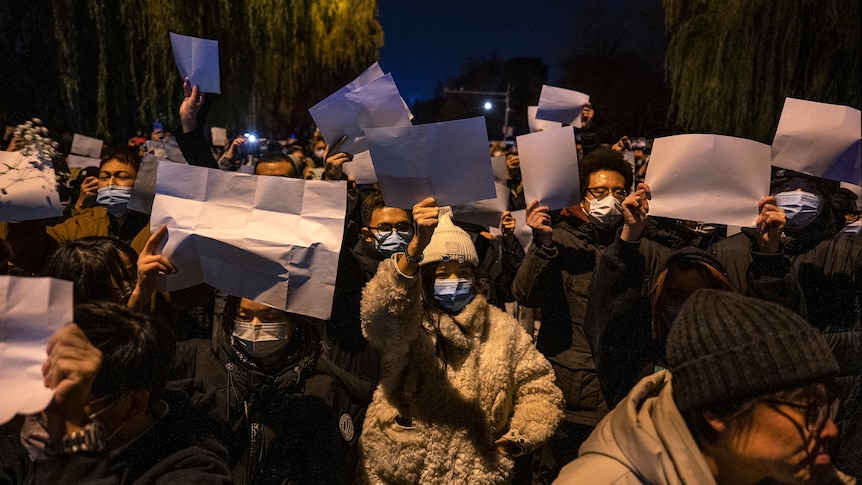 crowd of people wearing face masks holding up blank pieces of paper in protest