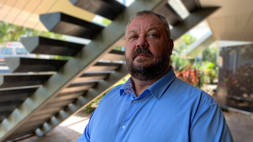 Greg Ireland wears a blue collared shirt, looking towards the camera, with stairs in the background.