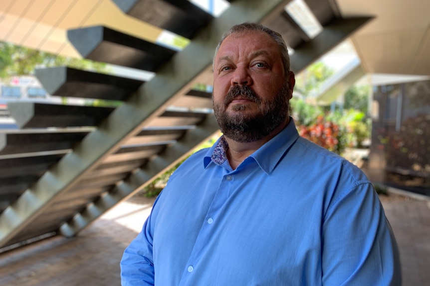 Greg Ireland wears a blue collared shirt, looking towards the camera, with stairs in the background.