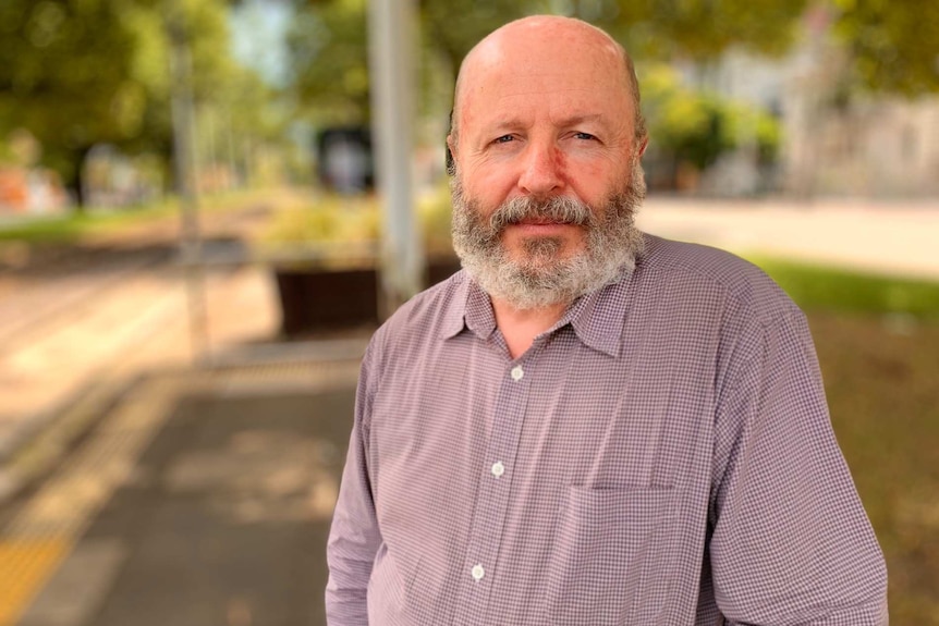 A middle-aged man with a beard wearing an open-neck shirt stares into the camera while waiting at a tram stop.
