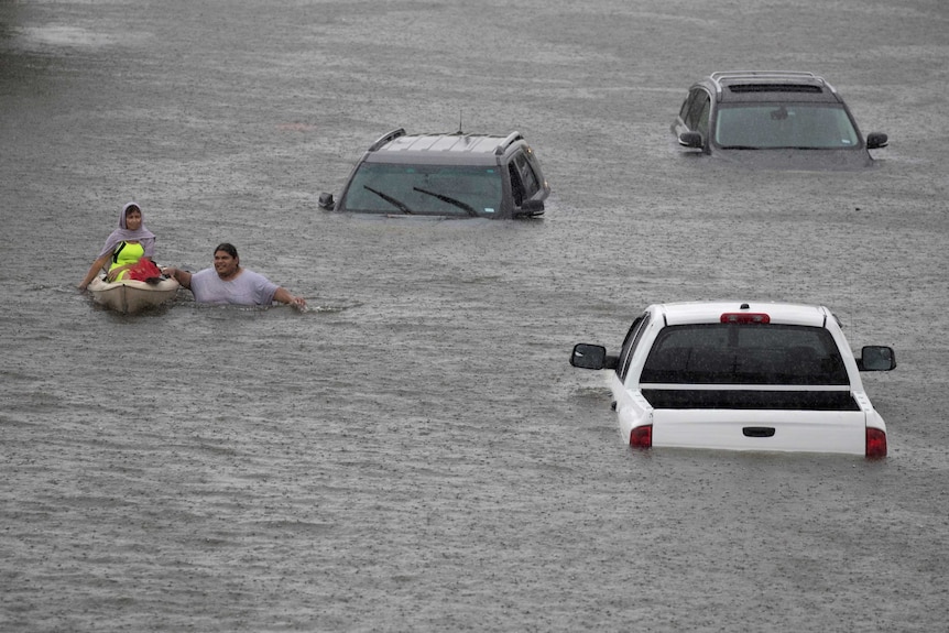 Two people, one in a canoe, make their way through floodwaters that reaches the windscreen on nearby cars.