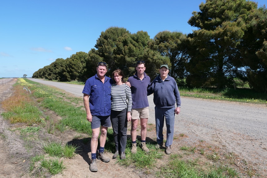 3 generations of men and a women stand next to a road. behind them a row of pine trees into the distance.