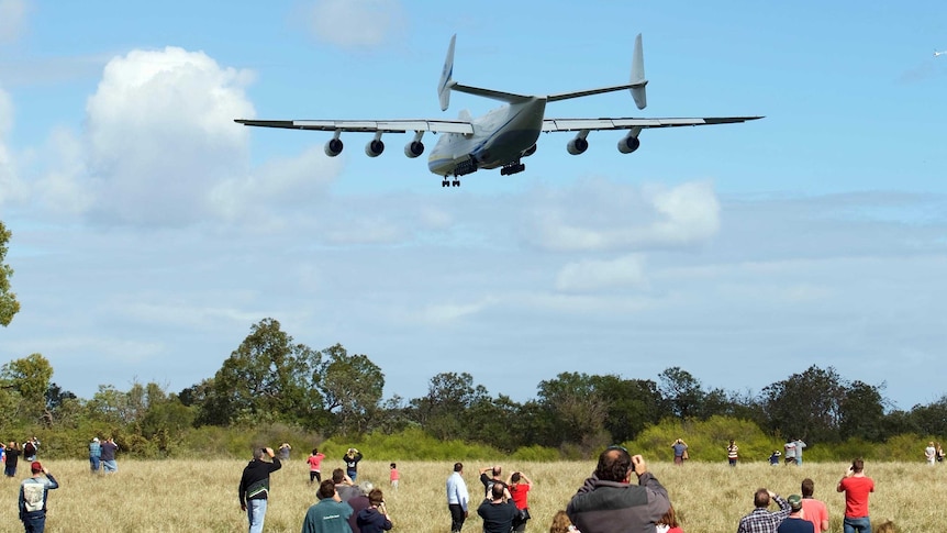Dozens of people standing in a grassy field watch and photograph the Antonov An-225 Mriya as it flies low overhead.