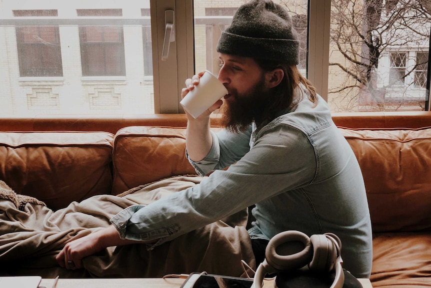 A man wearing a beanie sips from a paper cup while reclining on a couch, like he stayed over the night.