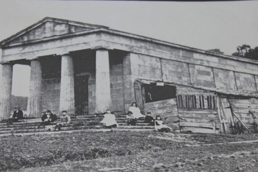 A black and white photo of a sandstone building with girls in 1800s clothing