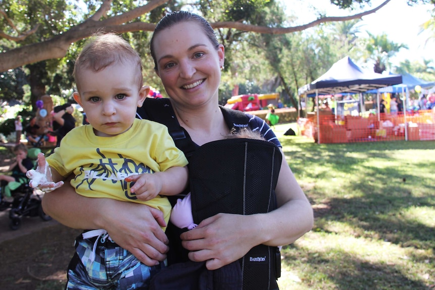 A woman holds her toddler and newborn baby in a park.