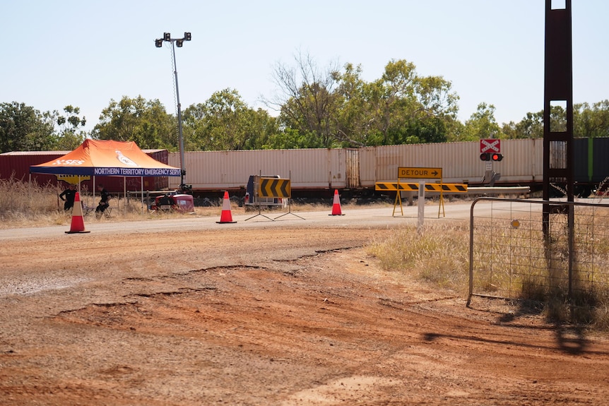 A train sits idle on a track crossing with a road closure sign in the foreground next to a police tent