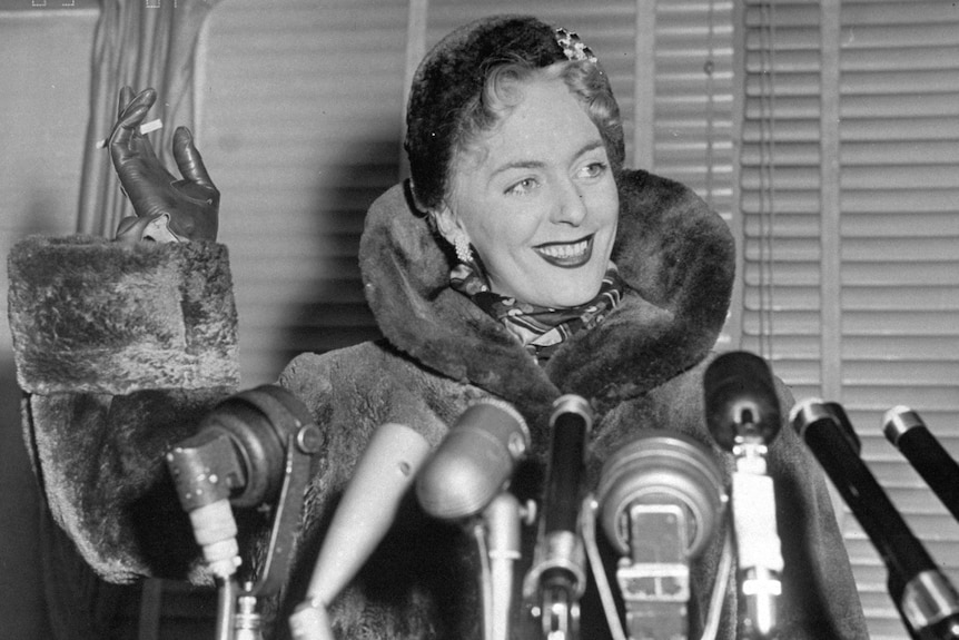 Black and white photo of Christine Jorgensen waving cigarette and smiling widely before multiple microphones and paparazzi.