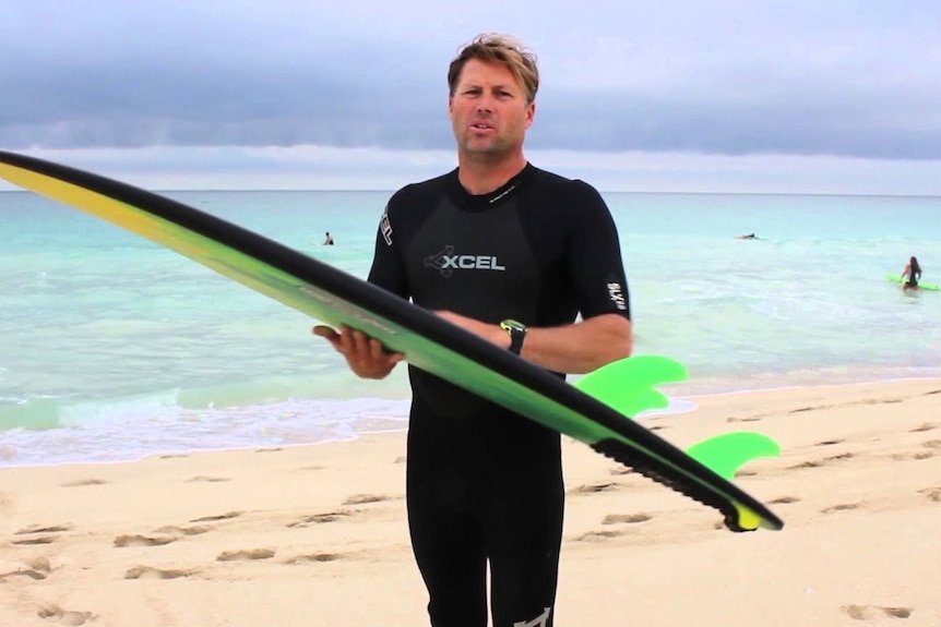 He stands in a wetsuit on a beach, holding a surfboard