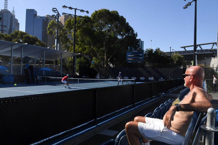 A spectator with his shirt off relaxes in the sun while watching a practice session at the Australian Open