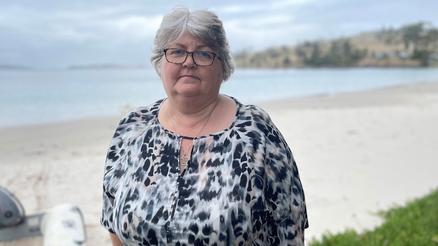 A woman with silver hair and glasses stands in front of a beach