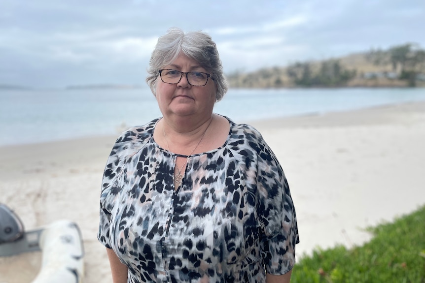 A woman with silver hair and glasses stands in front of a beach