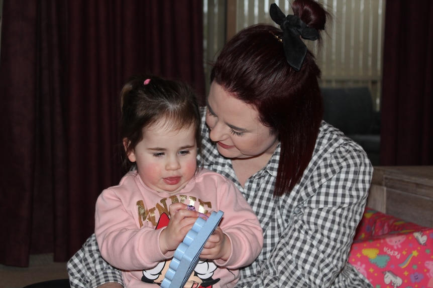 A woman with dark hair partially tied up with ribbon looks at toddler in lap, playing with toy. Woman wears black, white check.