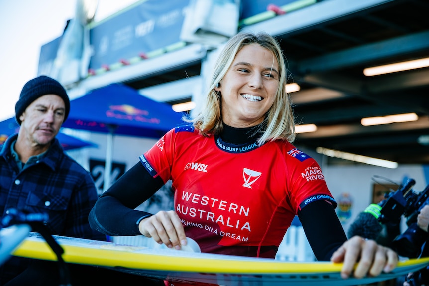 A surfer with blonde shoulder length hair smiles while holding her surfboard.