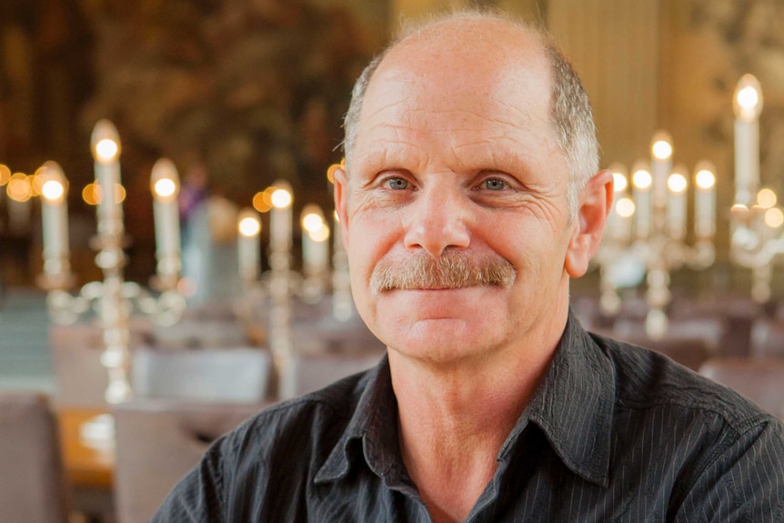 A balding man in his sixties poses for a portrait with a table of candelabras behind him.
