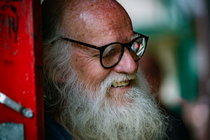 A close up shot of an older man with a white beard and dark glasses smiles and looks down and to the right.