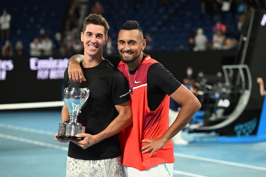 Tennis players Thanasi Kokkinakis (left) and Nick Kyrgios smile at the camera while Kokkinakis holds a trophy on court.