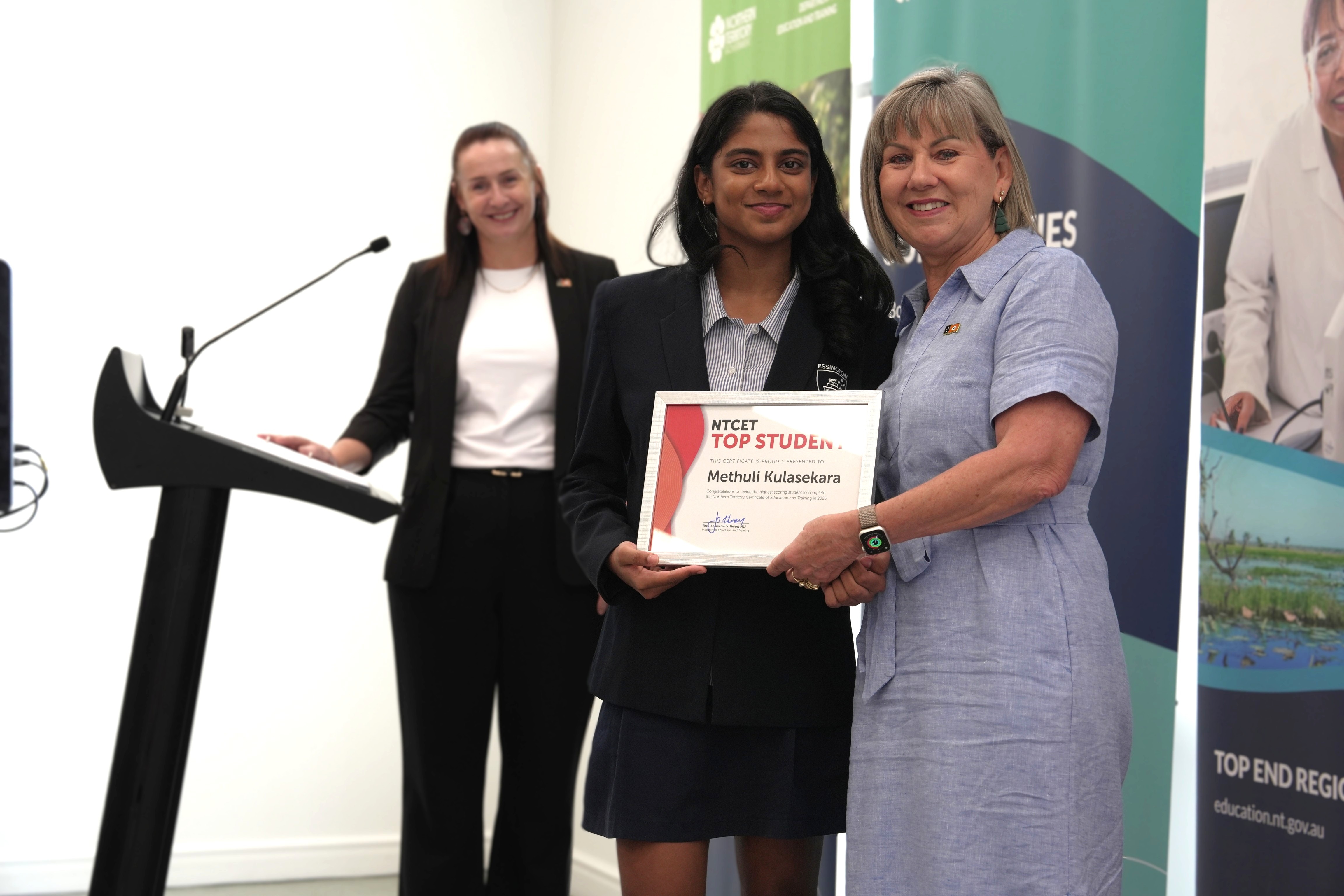 Un estudiante sostiene un certificado en la mano y posa con una mujer; al fondo, otra mujer sentada ante un atril.