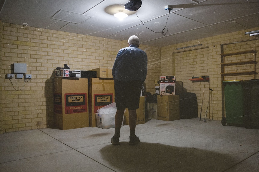 Dan stands in his dimly lit garage with cardboard boxes stacked along the wall
