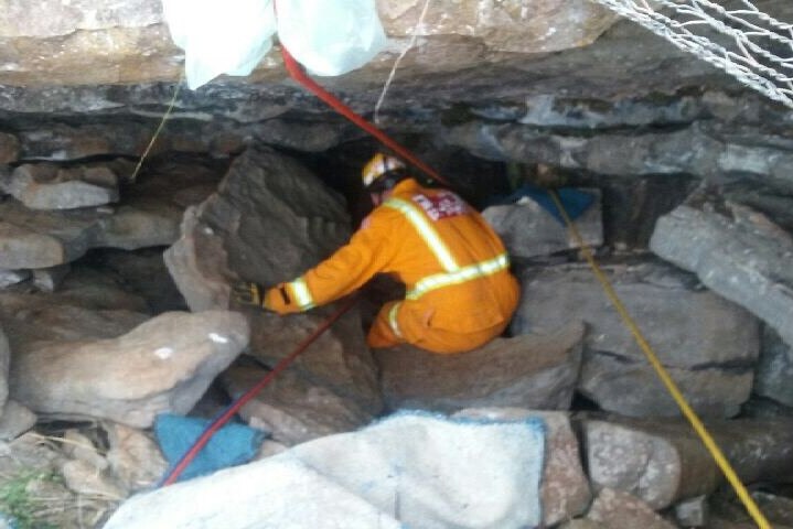 A person in an orange CFA uniform descends into a small cave with the help of ropes.