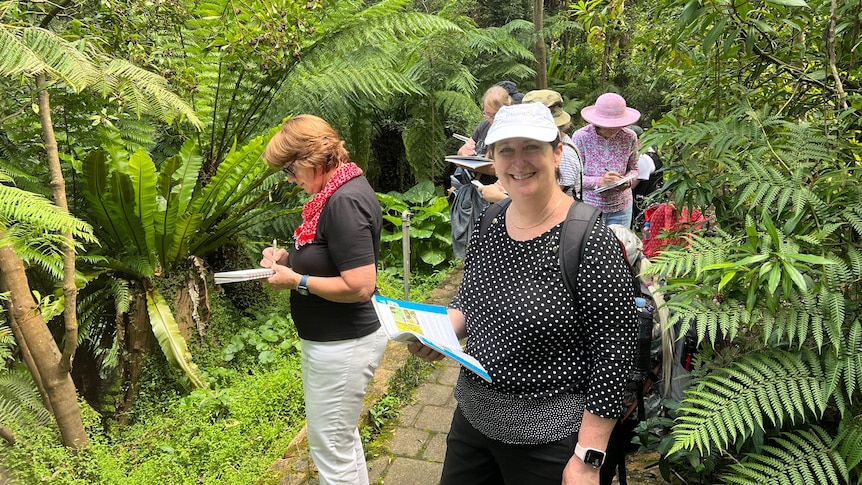 a woman is in a dark top and hat looking at the camera, holding a notebook. there are people and ferns in the background
