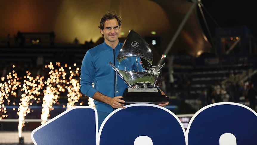 A man holds a trophy standing in front of a big number 100, with fireworks in the background.