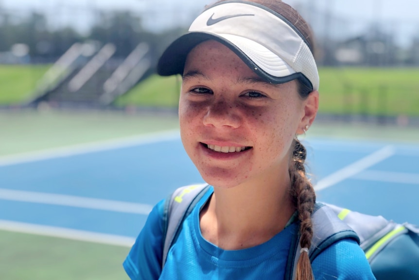 A close-up of a smiling Milly on a tennis court, wearing a sun visor, hair tn a pigtail, a backpack on shoulder.