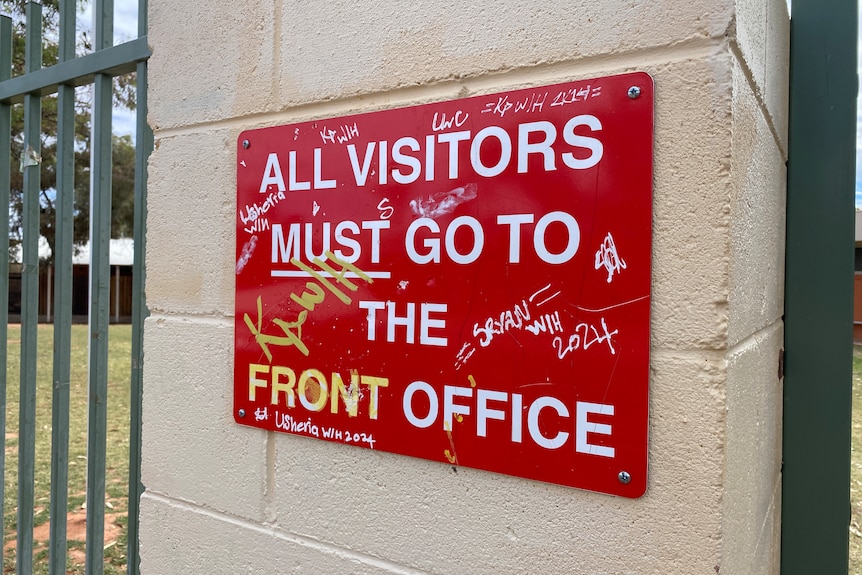 A red, graffitied sign on a school fence reads "all visitors must go to the front office".