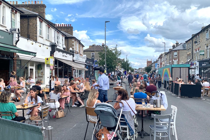 People sit gathered at dining tables set up on a street with buildings in the background.