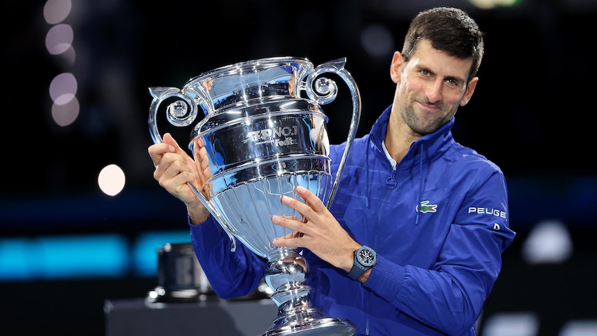 A male tennis player holds a trophy to his right as he poses for photographs