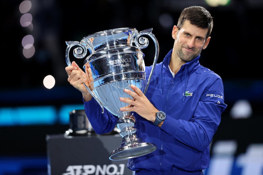 A male tennis player holds a trophy to his right as he poses for photographs