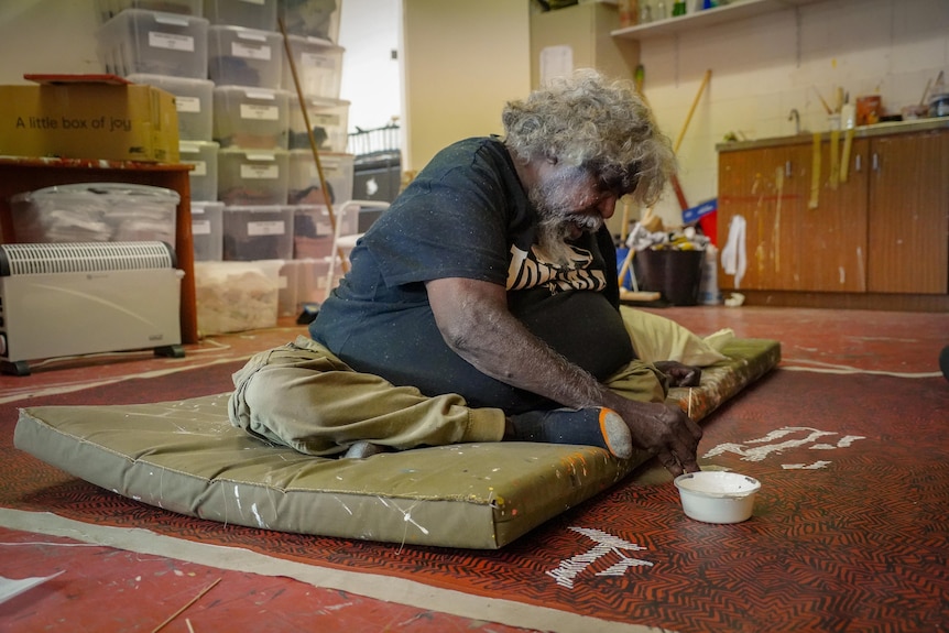 An Indigenous man with grey curly hair sits on the ground on a cushion, painting a large canvas. He is in an art studio setting.