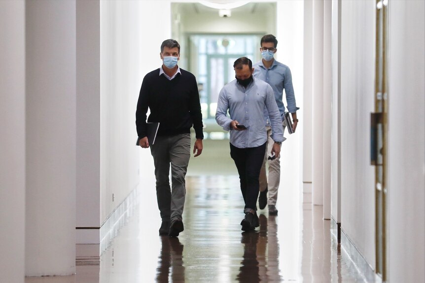 Three men walk along a hallway toward the camera.