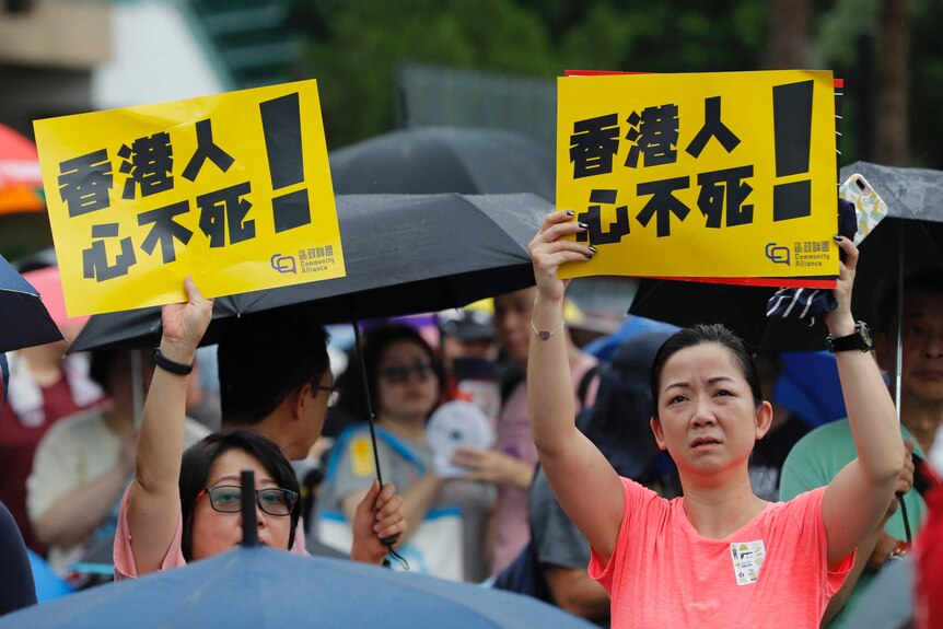 Two women in orange shirts hold a yellow sign that reads "Hongkongers won't give up!"