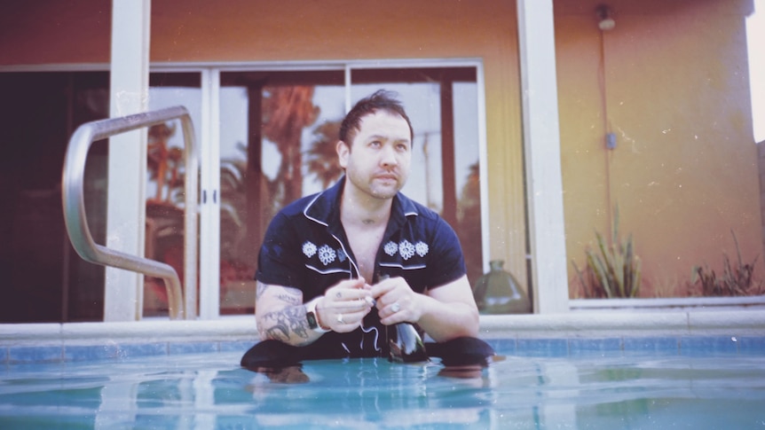 Unknown Mortal Orchestra's Ruban Nielson stands waist-high in a swimming pool holding a cigarette and wine bottle