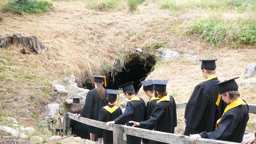 Students wearing black gowns and mortar boards walk down a stairway into a cave.