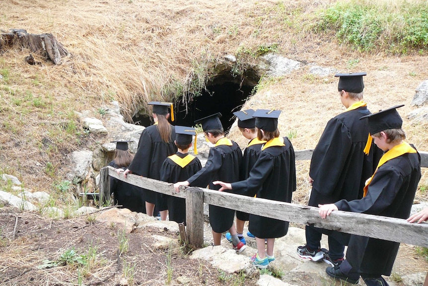 Students wearing black gowns and mortar boards walk down a stairway into a cave.