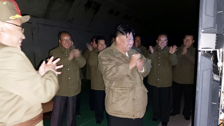 Kim Jong Un laughs and claps his hands surrounded by other men in military jackets. 