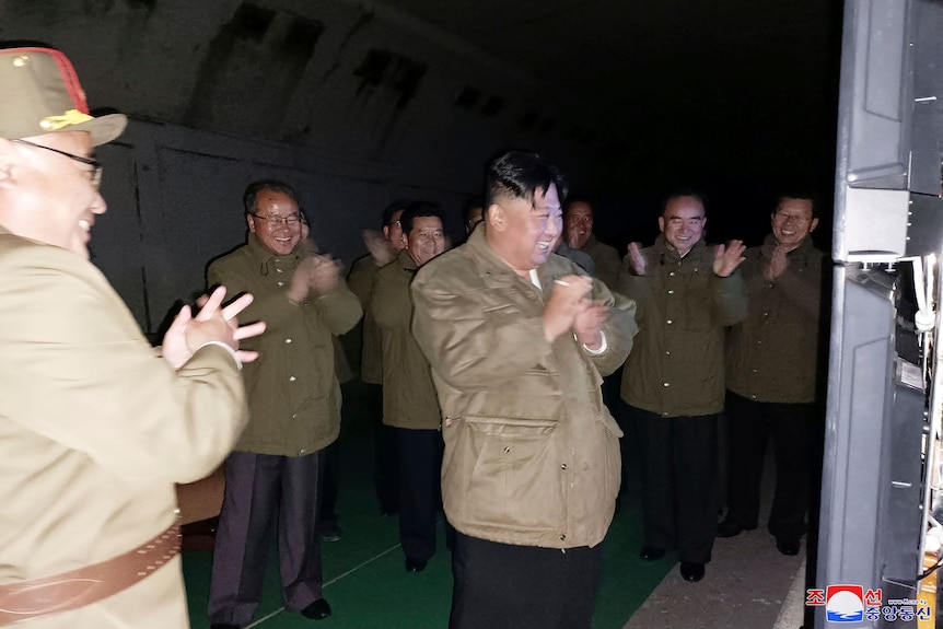 Kim Jong-un râde și bate din palme, înconjurat de alți bărbați în jachete militare. 