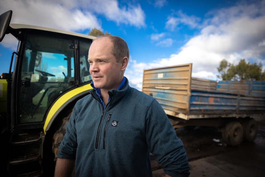 A man in workwear stands looking to the left of frame, with a tractor behind him 