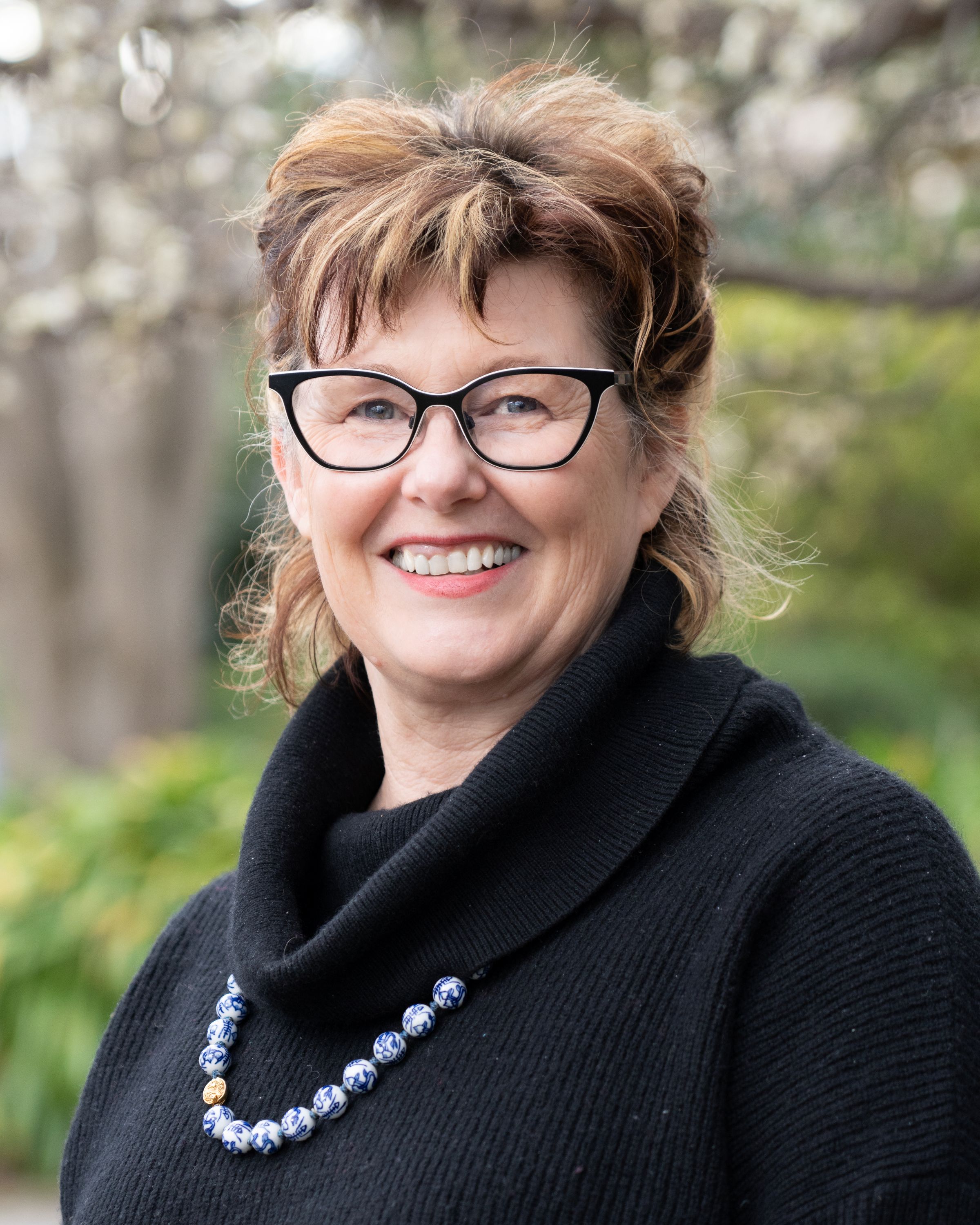 A woman with short brown hair and glasses, wearing a black cowl-neck top and a necklace
