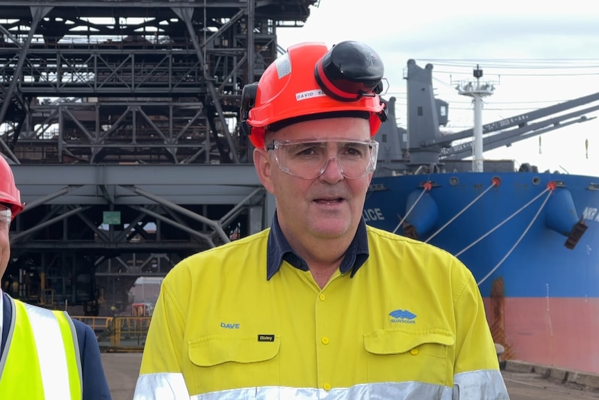 Man wearing hivis and a hard had with a ship behind 