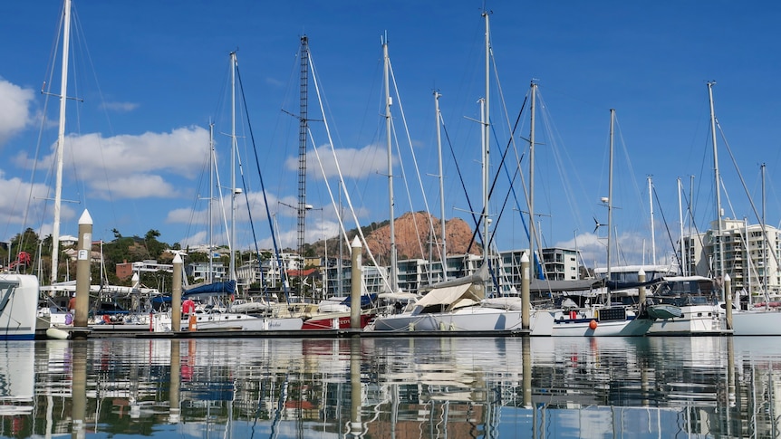 Large boats with tall masts sit in a marina with the still seawater just below. There are tall buildings and a mountain behind.