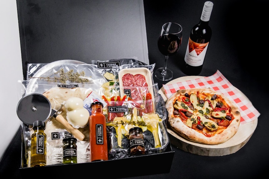 A box filled with ingredients to make pizzas, a full wine glass and a ready-made pizza.